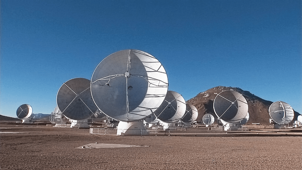 An image of the ALMA telescope array, with 9 or more large radio antennae pointed towards the camera atop a dry, arid mountain.