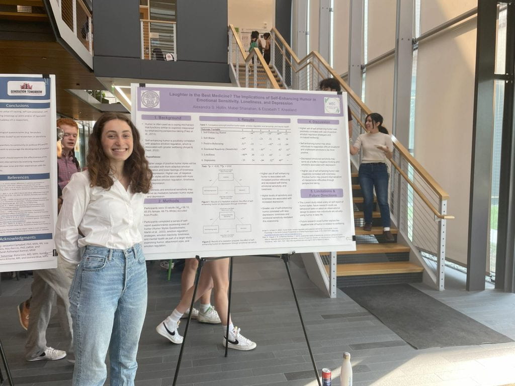 A woman with long, curly hair wears light-washed jeans and a white button-up shirt. She smiles in front of a scientific-style poster. A staircase is behind her, and people mill about in the background.