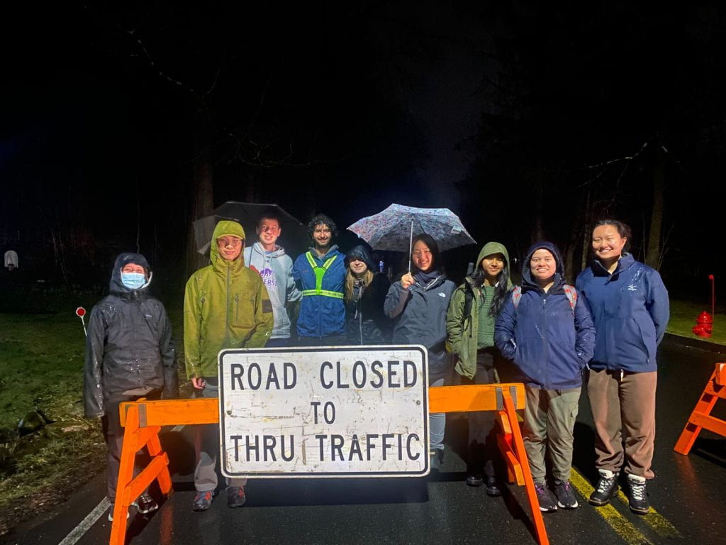 Nine students stand behind a "Road Closed to Thru Traffic" sign tacked to an orange barrier. They are decked out in rain gear and umbrellas. The darkness shrouds the scene behind them.
